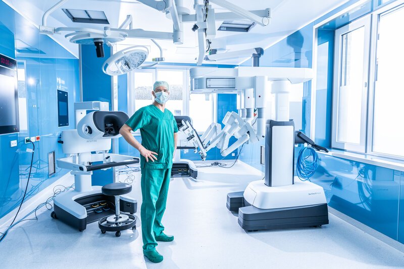 Chirurg im OP-Saal neben einem robotergestützten Operationssystem und weiteren medizinischen Geräten.